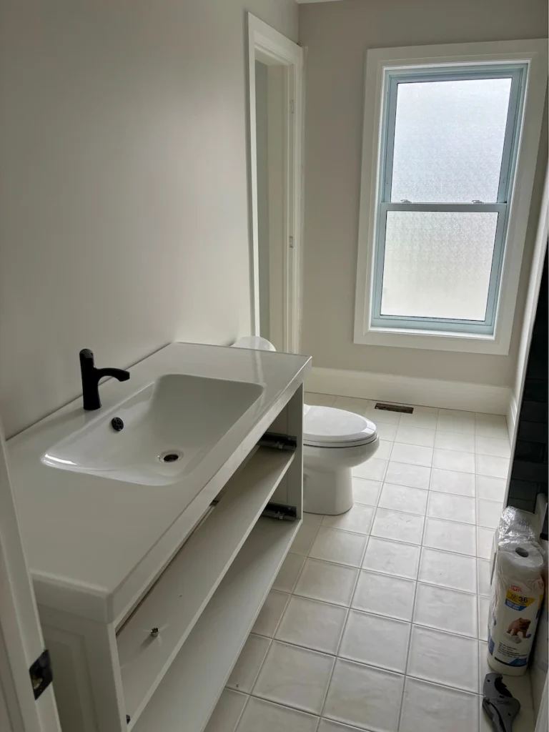 Bathroom with a white vanity and sink, black faucet, tiled floor, and a toilet near a window.