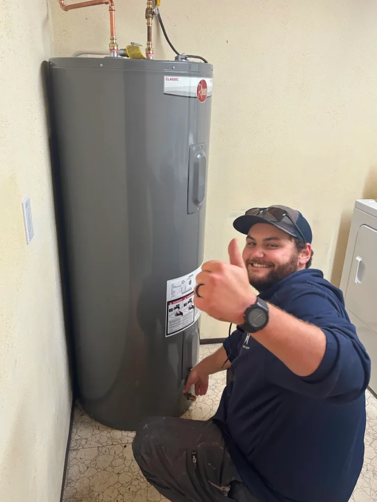 Technician kneeling beside a Rheem water heater and giving a thumbs up gesture.
