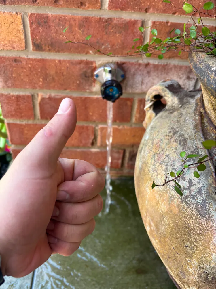 Outdoor hose bib mounted on a brick wall with water flowing from the spout and a hand giving a thumbs up in the foreground.